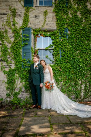 Wedding couple poses against ivy-covered stone wall with blue shuttered windows.