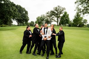 Groomsmen in black tuxedos posing playfully on green lawn.