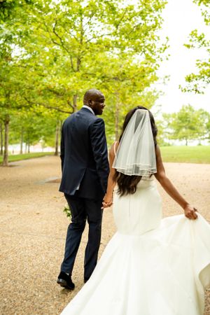 Wedding couple strolls together under a canopy of green trees.