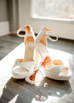 Elegant white beaded bridal heels with ankle straps sitting in natural window light on dark surface.