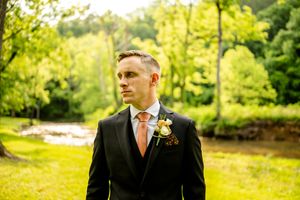 A groom in a black suit with orange tie poses in a sunlit garden setting.