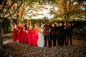 Wedding party in red and black attire poses together under autumn trees.
