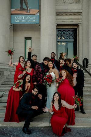 Joyful wedding party celebrating on museum steps wearing red dresses and black suits.