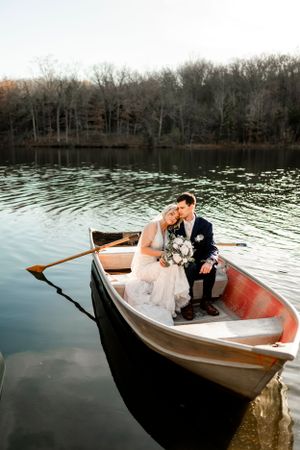 Newlyweds share an intimate moment in a wooden rowboat on a peaceful lake.