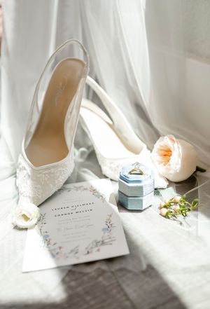 Wedding details including white lace shoes, invitation and ring box in soft natural light.