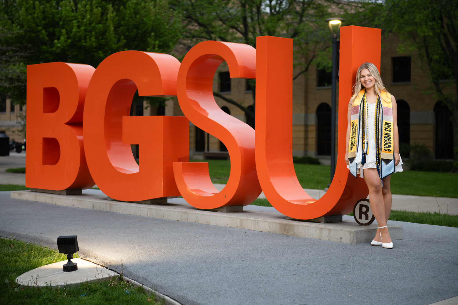 A smiling graduate poses beside large orange BGSU letters on campus, wearing a decorated graduation stole.