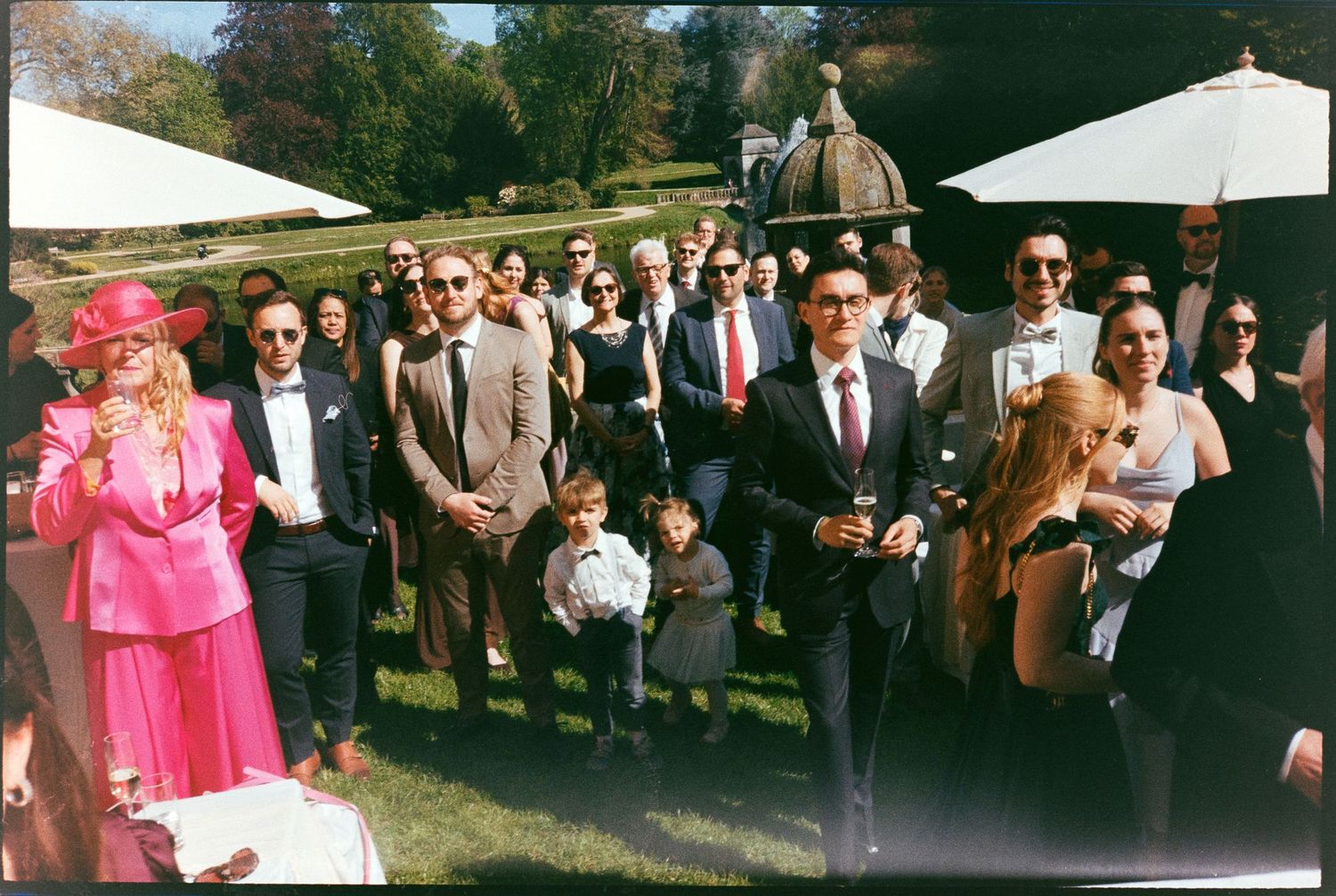Elegant outdoor garden party with guests in formal attire, including a woman in pink hat and suit, on a sunny day.