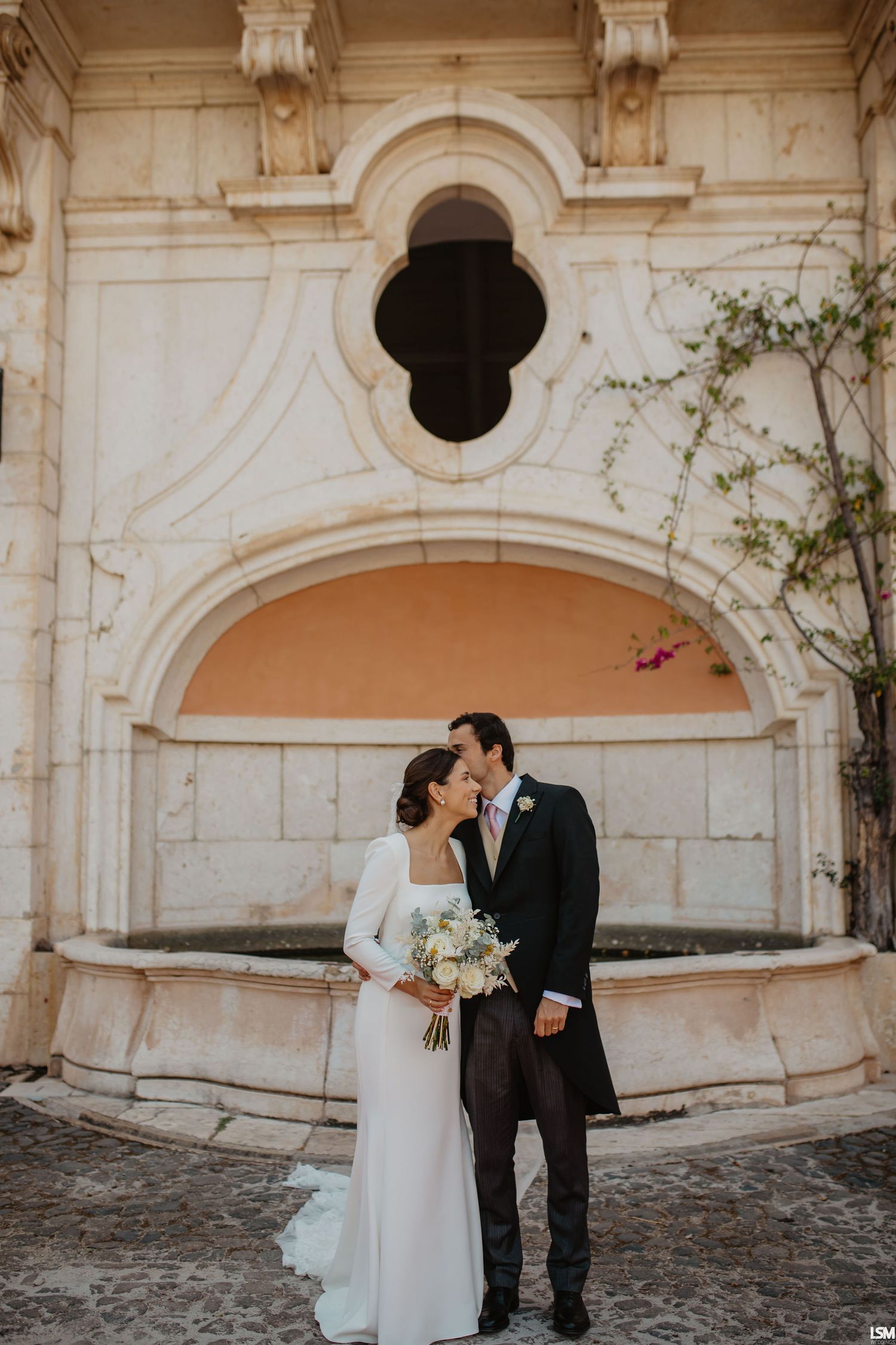 Bride and groom share a romantic kiss in front of an ornate stone fountain arch, bride holding a white bouquet.