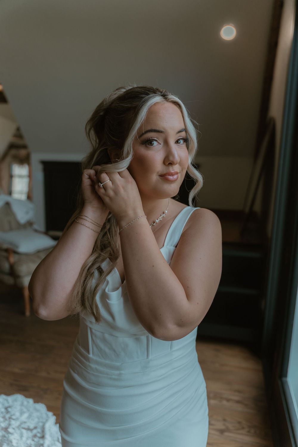 Bride in white gown putting on earrings in dimly lit room.