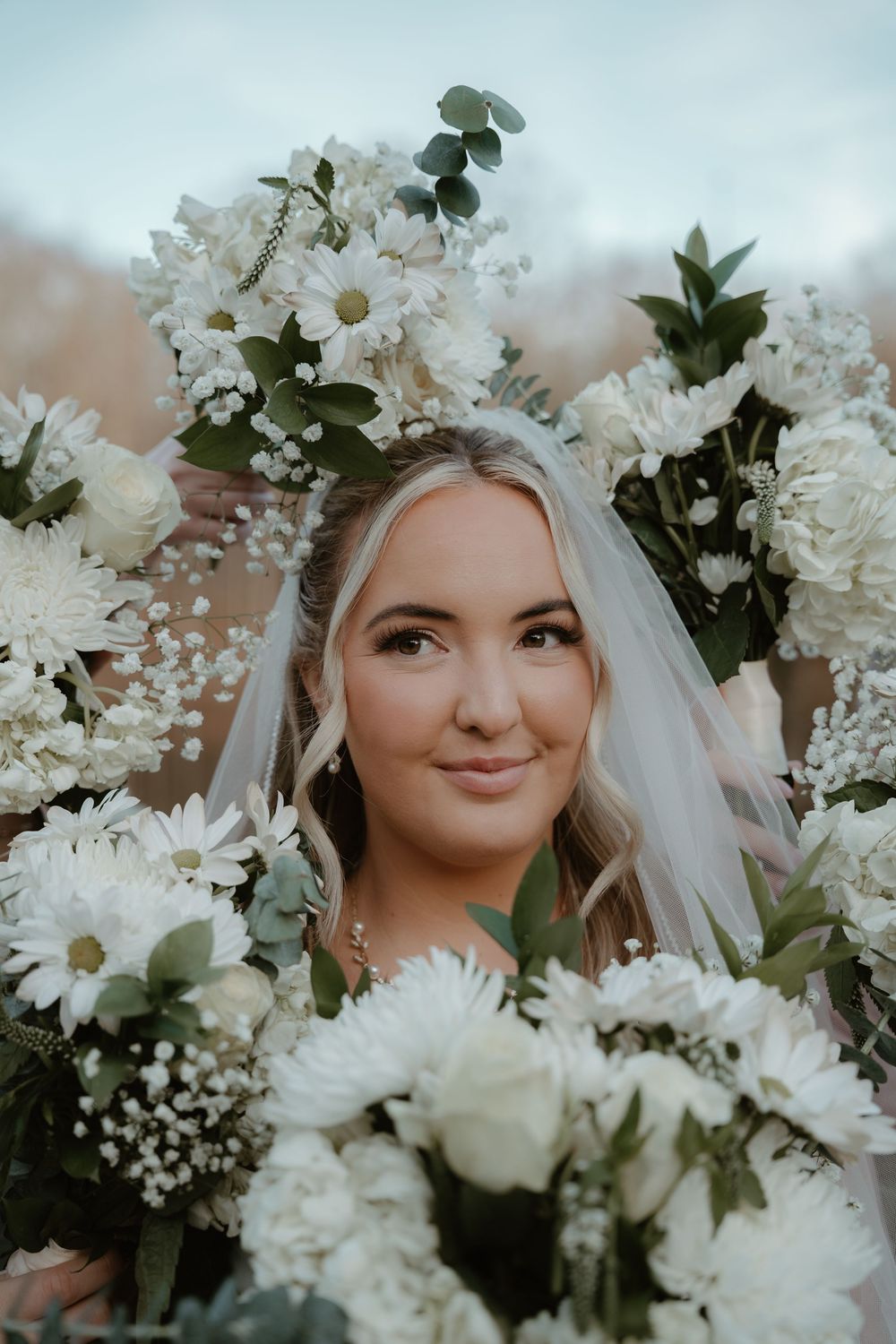 Bride surrounded by white flowers and greenery wearing a veil on her wedding day.