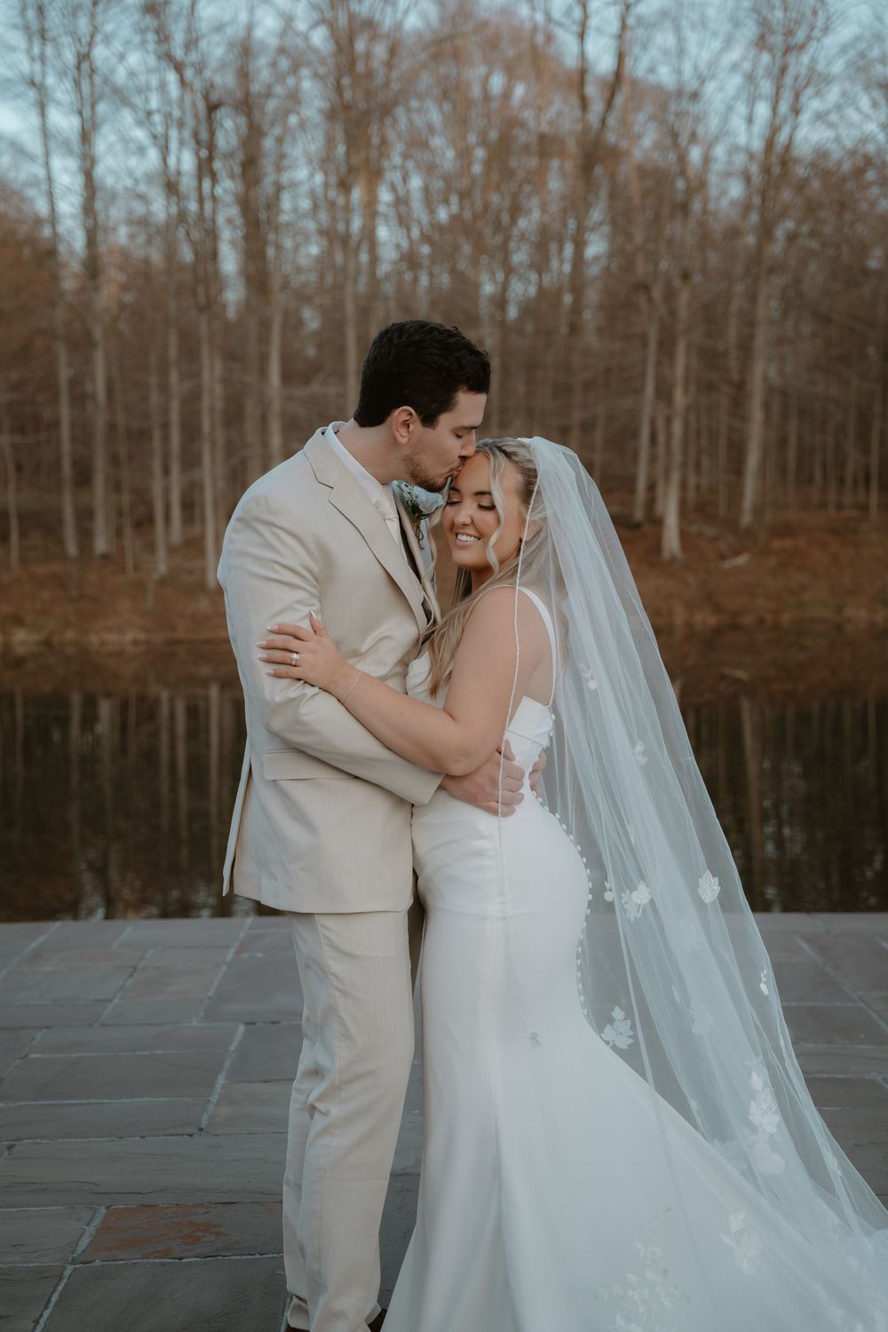Bride and groom sharing a romantic embrace by a serene lake in autumn surroundings.