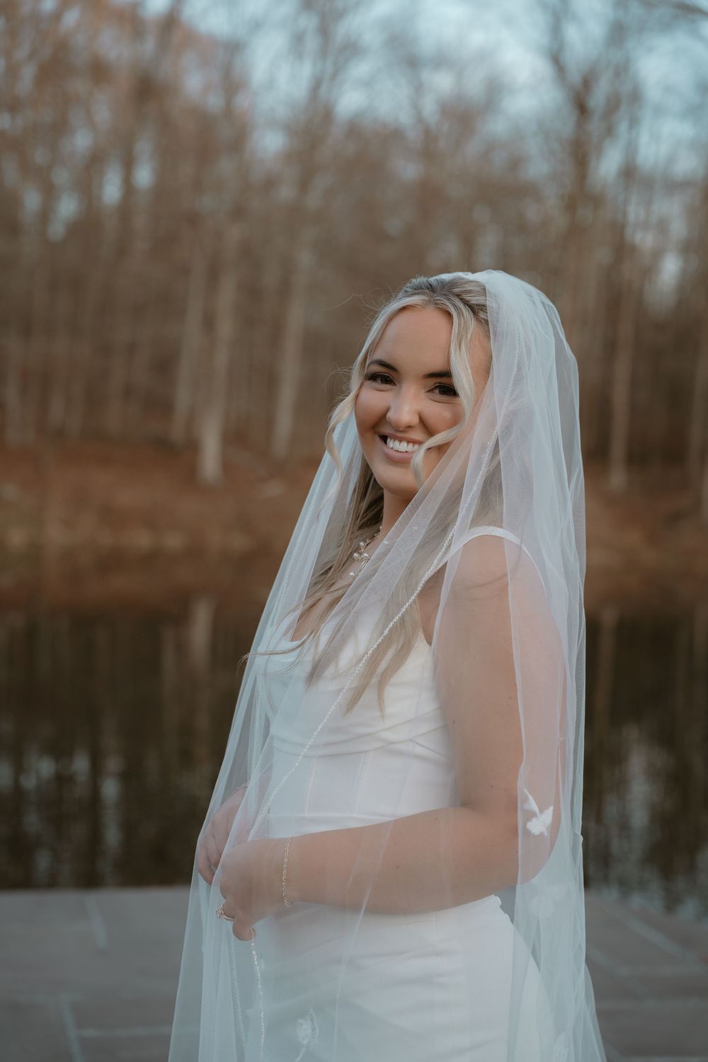 Smiling bride in a white gown and veil standing near a reflective lake at dusk.