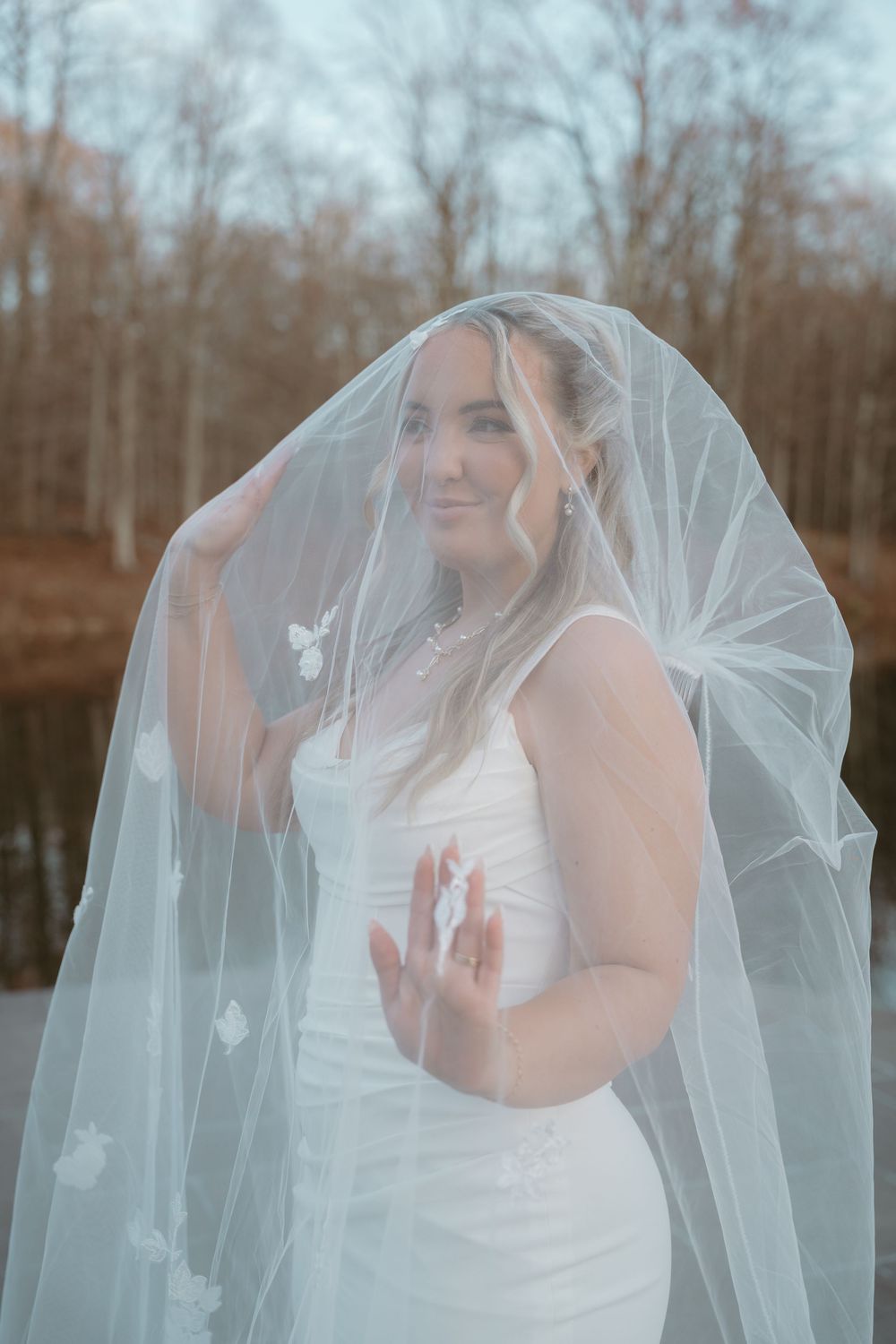 Bride lifting her floral embroidered veil with a serene smile at an outdoor fall wedding.