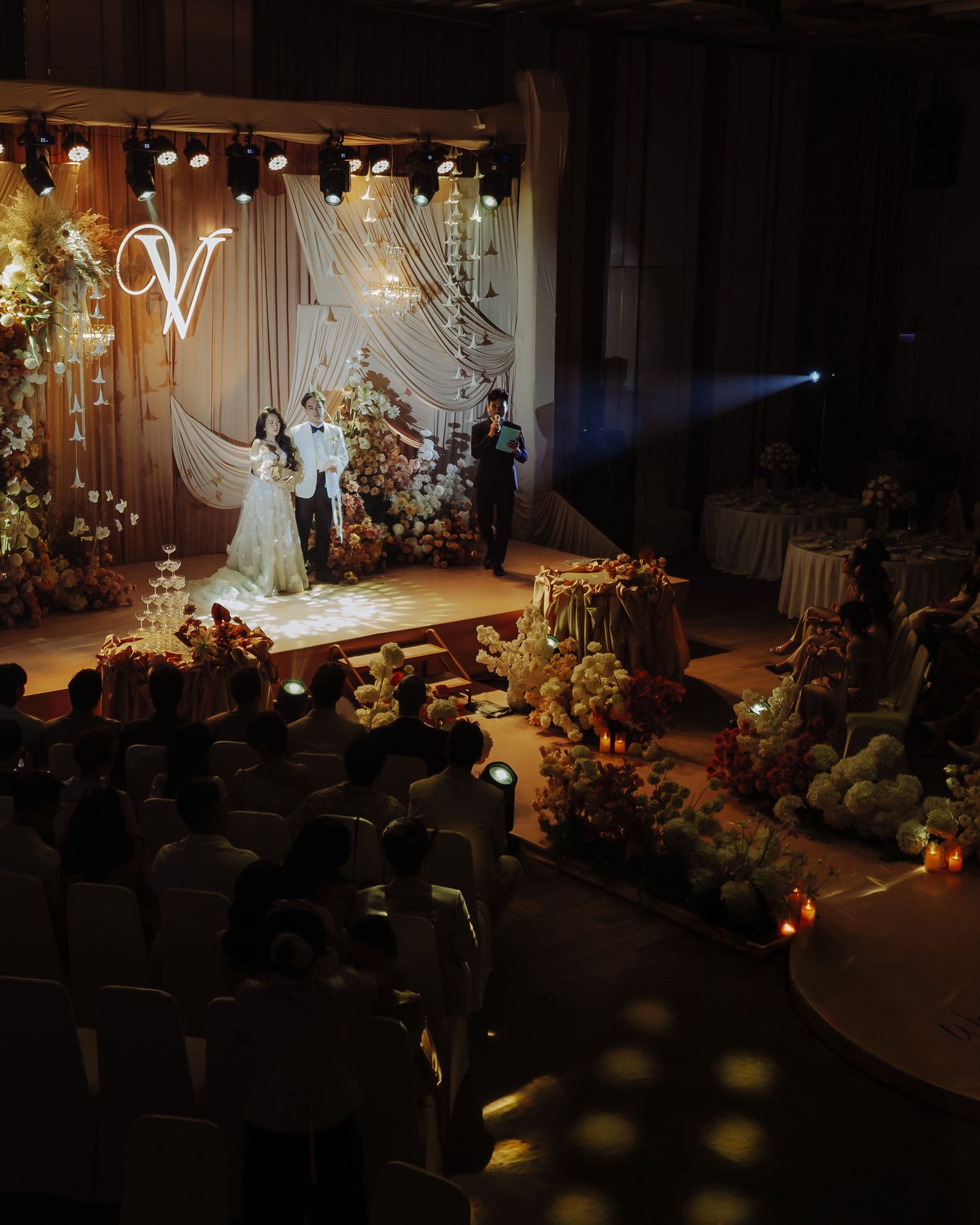 Dramatic indoor wedding reception with glowing stage, floral decor, and monogram backdrop.