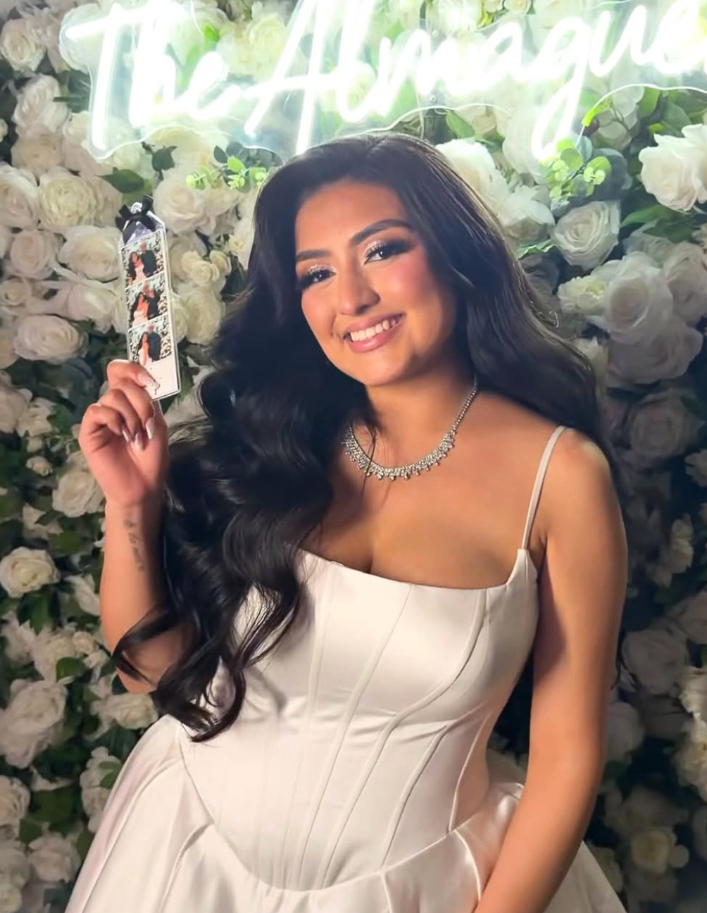 Young woman in white dress holding a photo strip, wearing a diamond necklace, posing in front of a white floral wall.