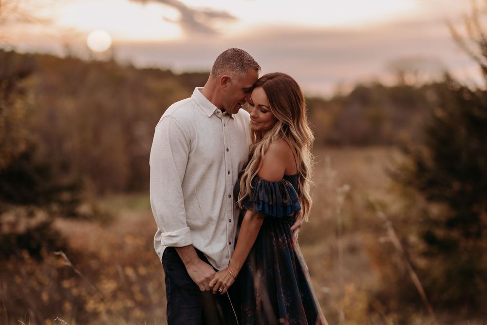 Couple embracing at golden hour sunset in a field, woman in navy off-shoulder dress, romantic outdoor engagement photo.