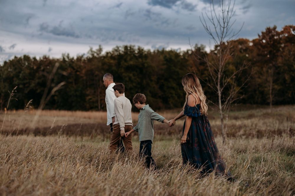 Family of four walking through autumn field at dusk, holding hands among tall golden grasses with trees in background.