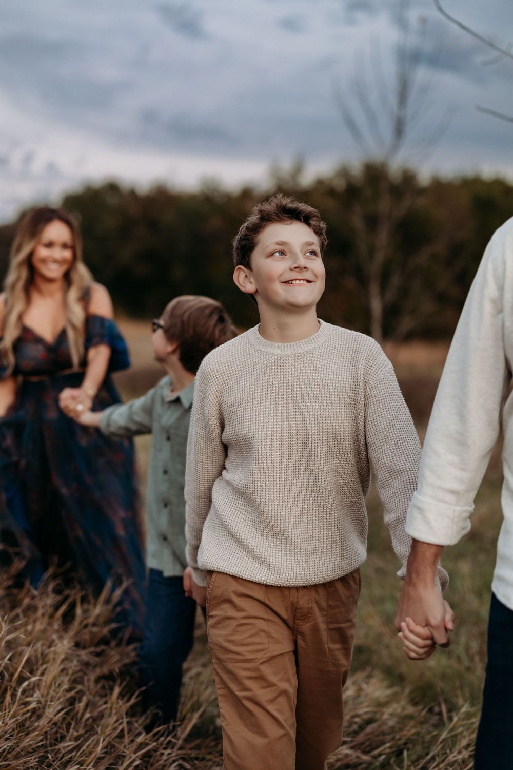 A smiling boy in a beige sweater holds hands with family while walking through a field at dusk.