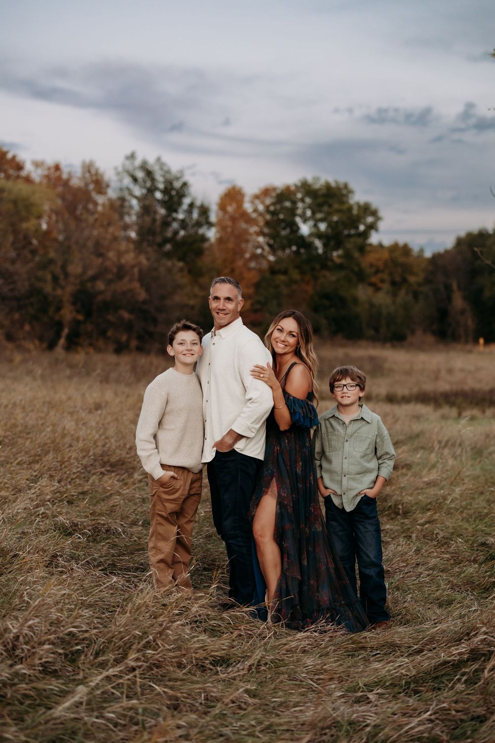 Family of four posing in an autumn field with colorful trees, wearing casual fall outfits during an outdoor photo session.