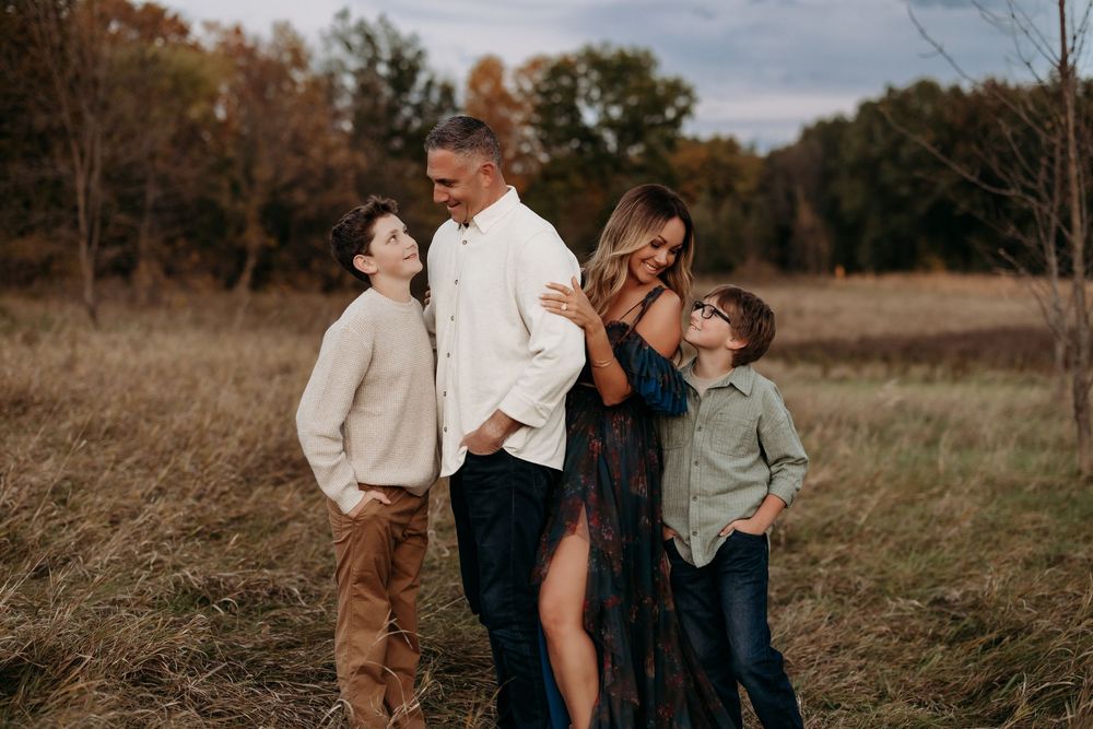 Family of four posing in autumn field, parents embracing while two boys stand beside them in warm fall colors.
