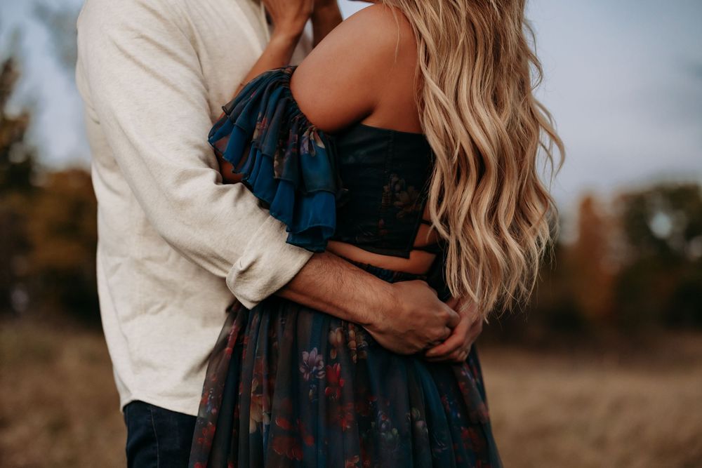 Couple embracing outdoors in autumn, woman in floral off-shoulder dress with long blonde hair, man in white shirt.