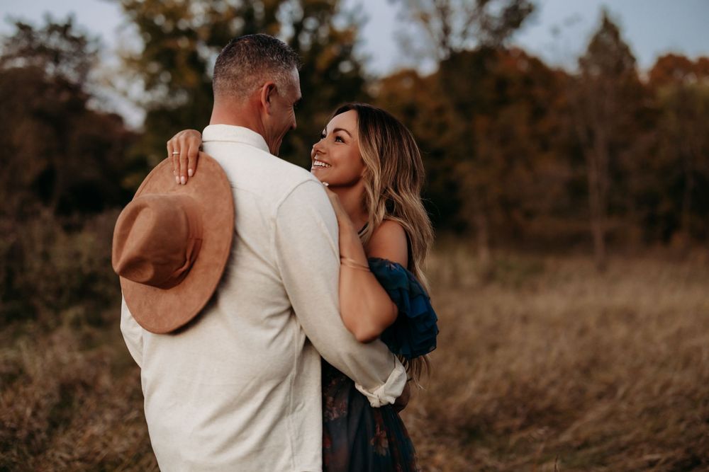 Couple embracing in autumn field, woman smiling in floral dress, man in white shirt holding tan hat.