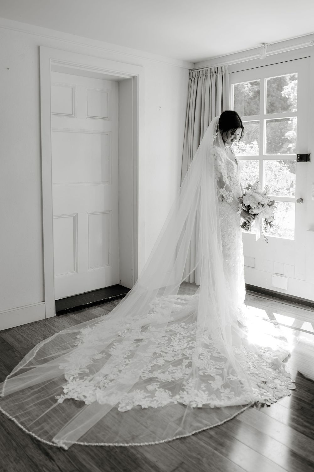 Bride in lace wedding gown with cathedral veil trailing on hardwood floor, holding bouquet in black and white photo.