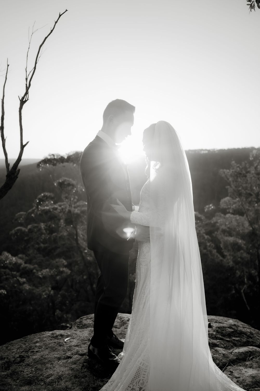 Black and white photo of bride and groom silhouetted against bright sun on rocky cliff with forest backdrop.