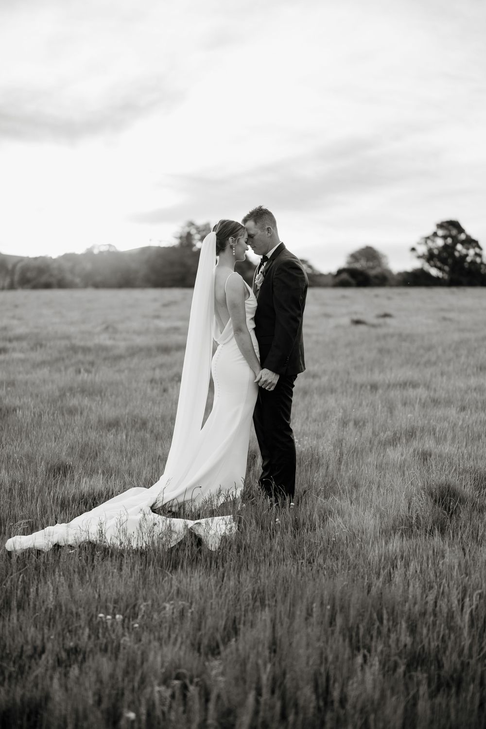 Black and white photo of bride and groom standing in a field, sharing an intimate moment in wedding attire.