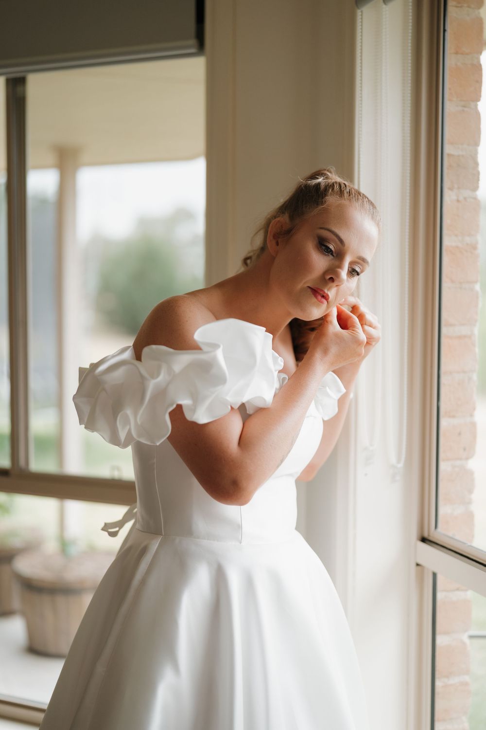 Bride in off-shoulder white wedding gown with ruffle sleeves stands by window, adjusting earring.