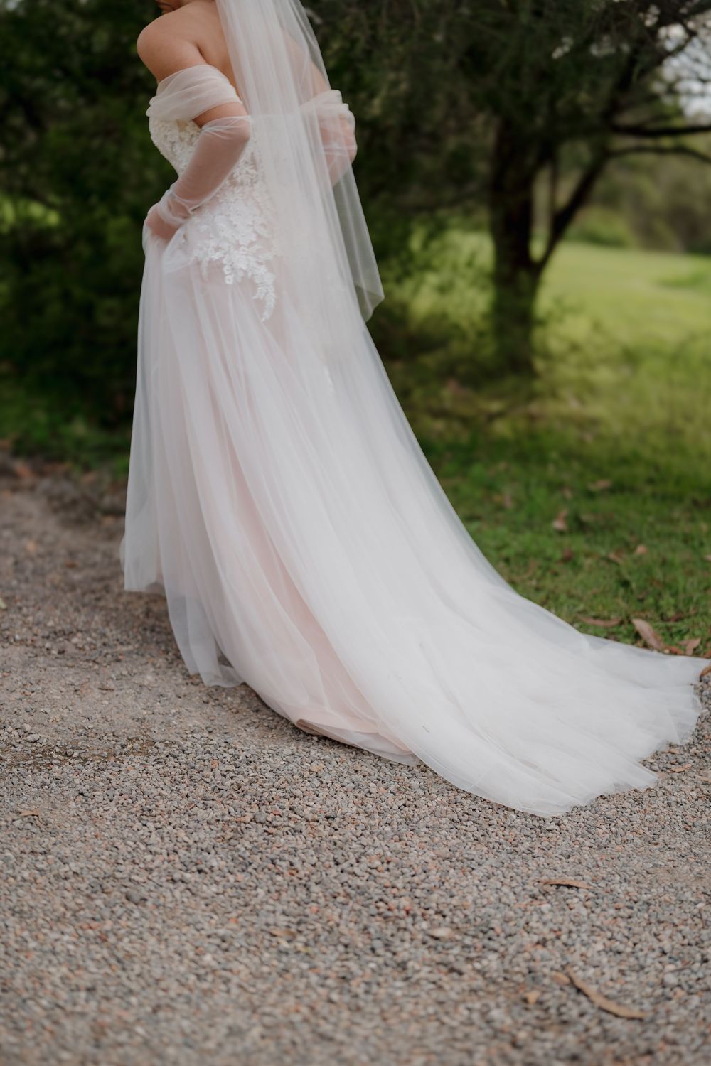 Bride in a flowing white tulle wedding gown with lace bodice and veil, standing on a gravel path outdoors.