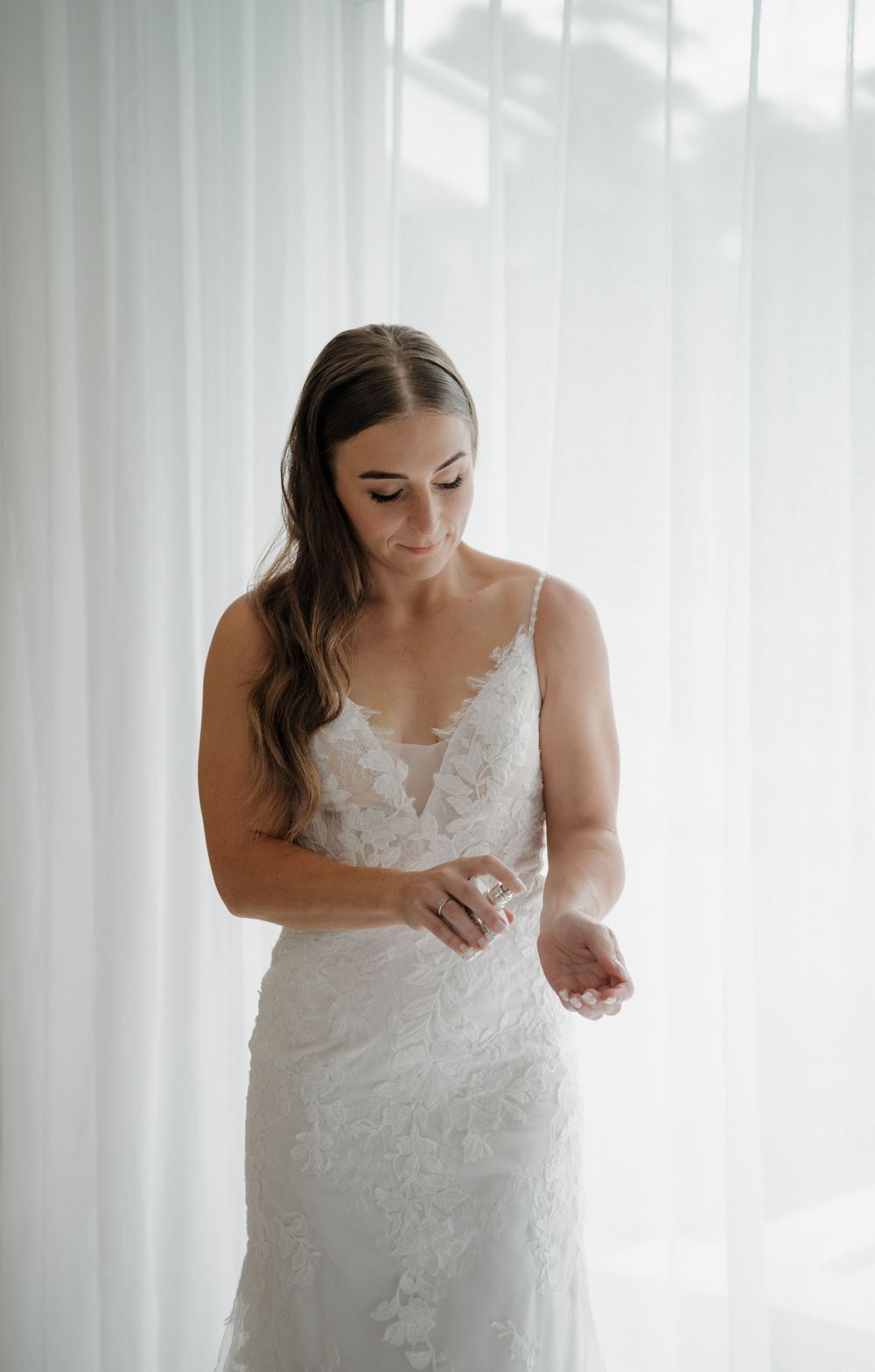 Bride in a lace spaghetti strap wedding gown applying perfume by a sheer white curtained window.