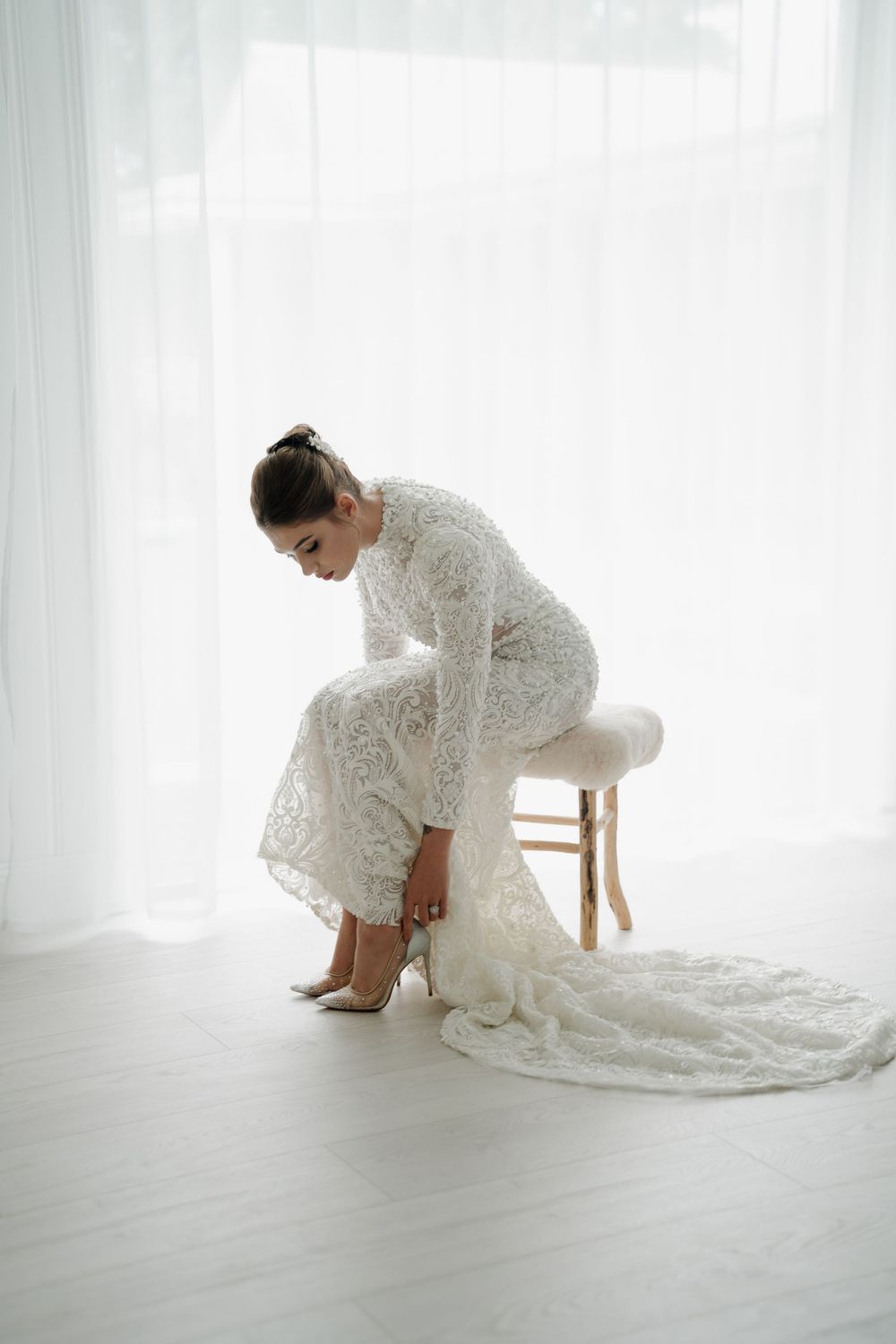 Bride in a long-sleeve lace wedding gown sits on a stool adjusting her heels before the ceremony.