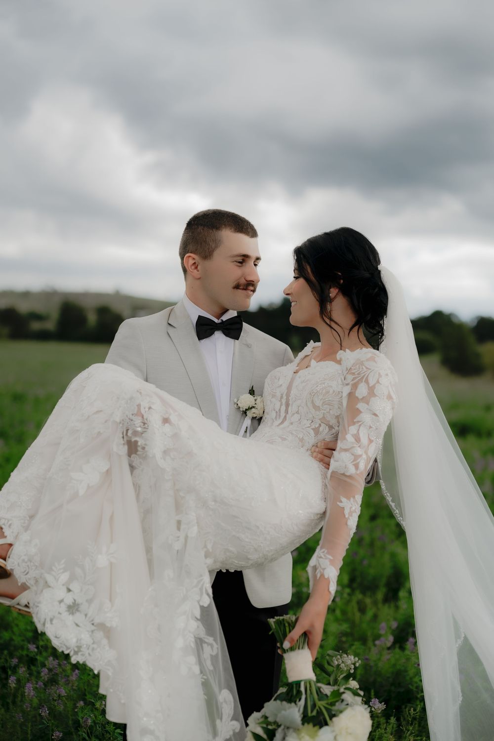 Groom in white tuxedo carries bride in lace wedding dress outdoors in green field under cloudy sky.