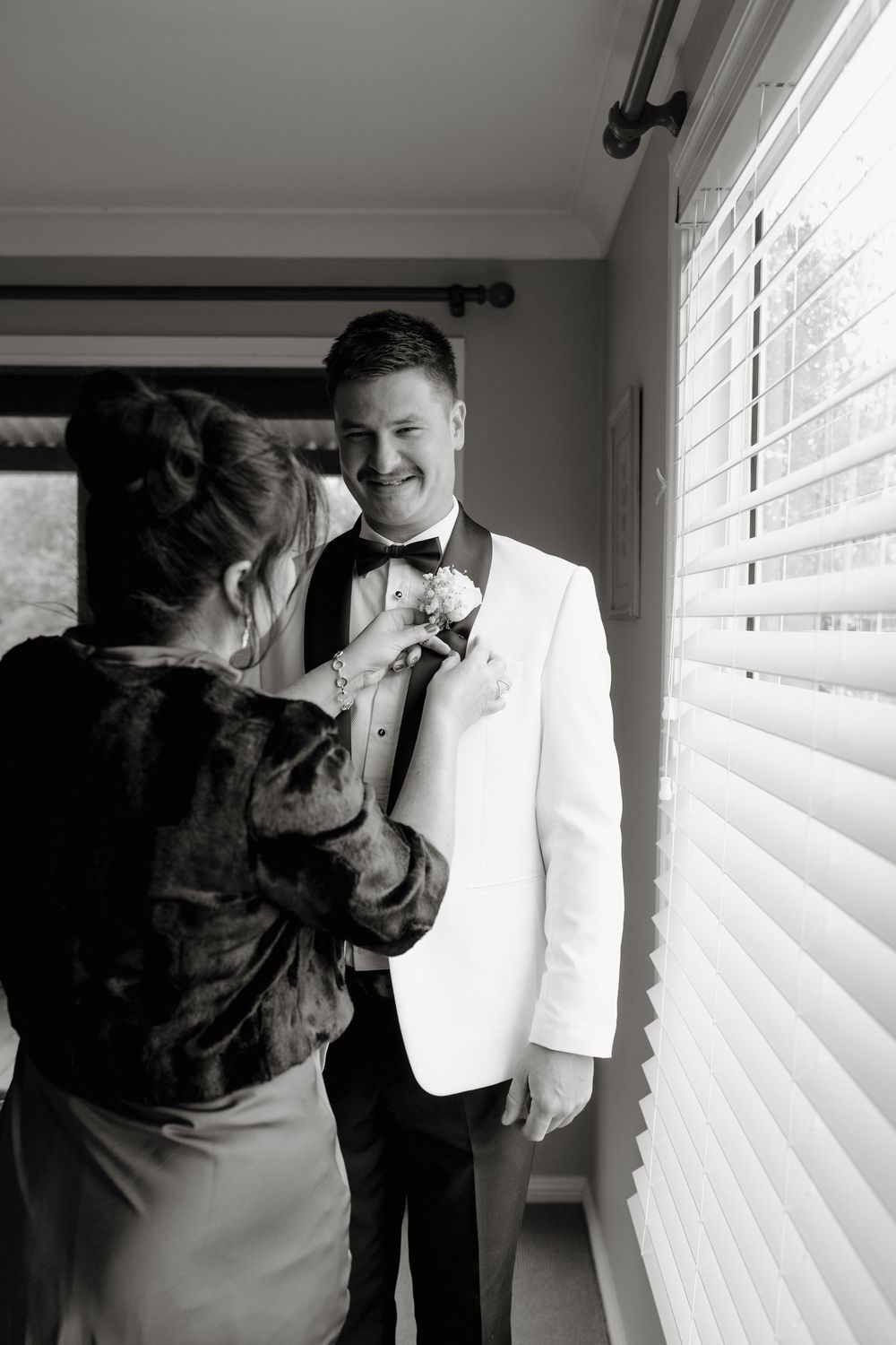 Black and white photo of a groom in white tuxedo smiling as a woman pins a boutonniere on his lapel.