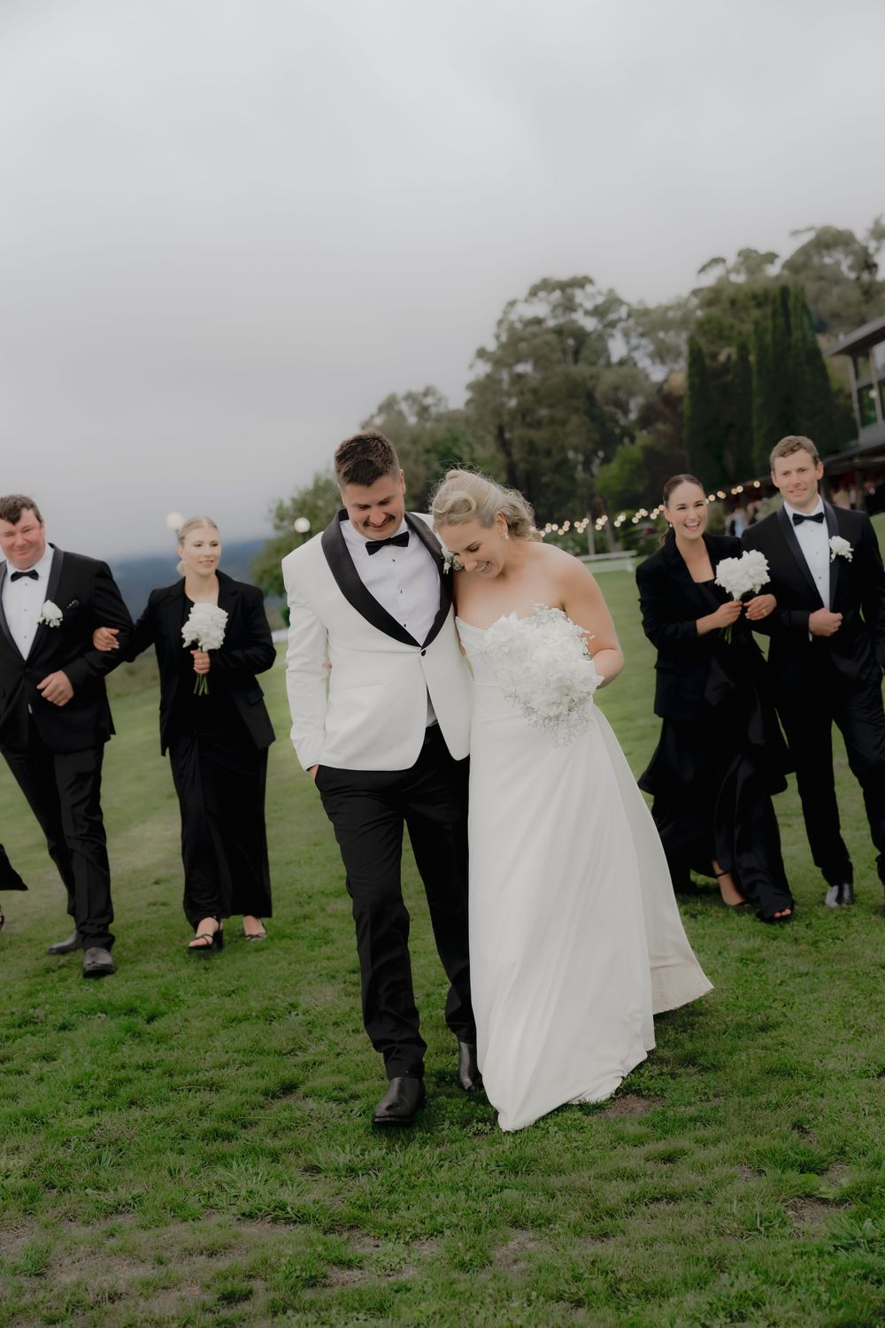 Bride and groom walk together on green lawn, bridal party in black attire follows behind at outdoor wedding ceremony.