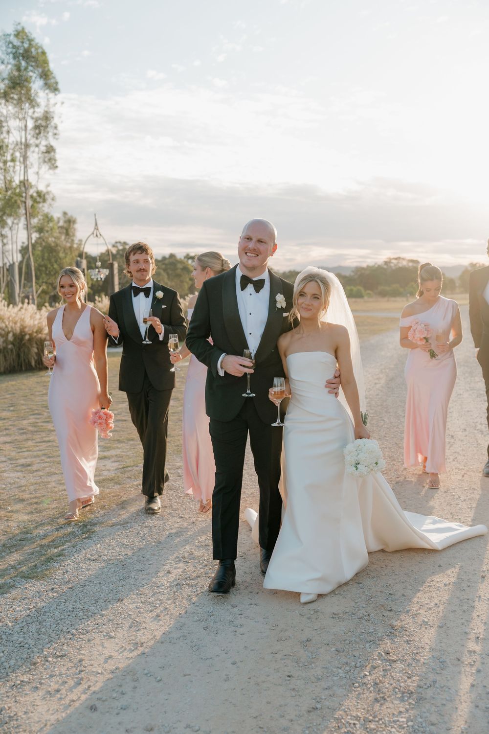 Wedding party walks on gravel path at golden hour, bride in white gown with bouquet, bridesmaids in pink dresses.