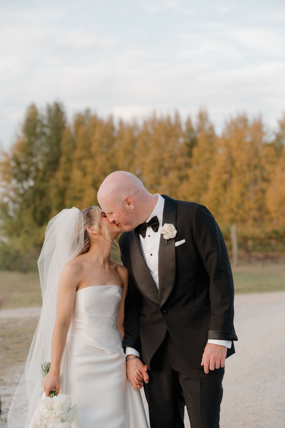 Bride and groom sharing a kiss on a gravel path with golden autumn trees in the background.