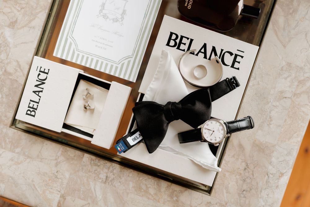Elegant groom accessories flat lay featuring BELANCE watch, black bow tie, cufflinks, ring, and wedding invitation on marble tray.