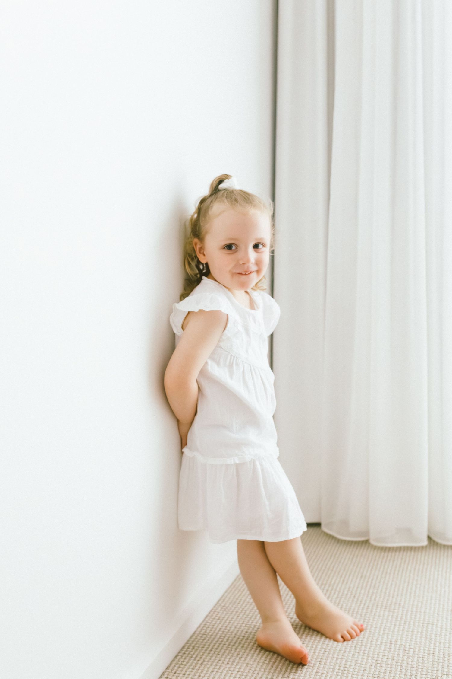 A young girl in a white dress and skirt leans against a white wall, smiling, with sheer curtains in the background.