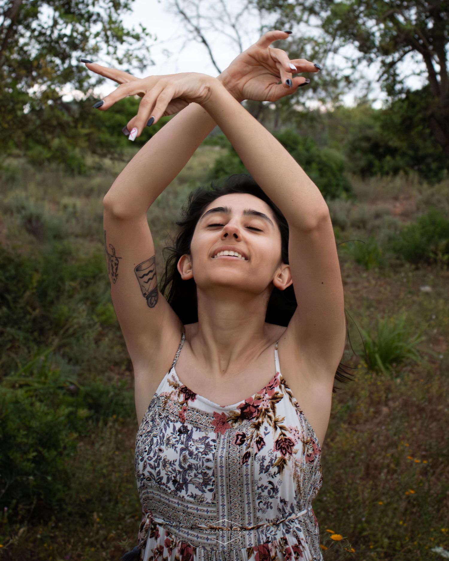 Woman in floral dress with arms raised overhead, smiling with eyes closed in a lush green outdoor setting.