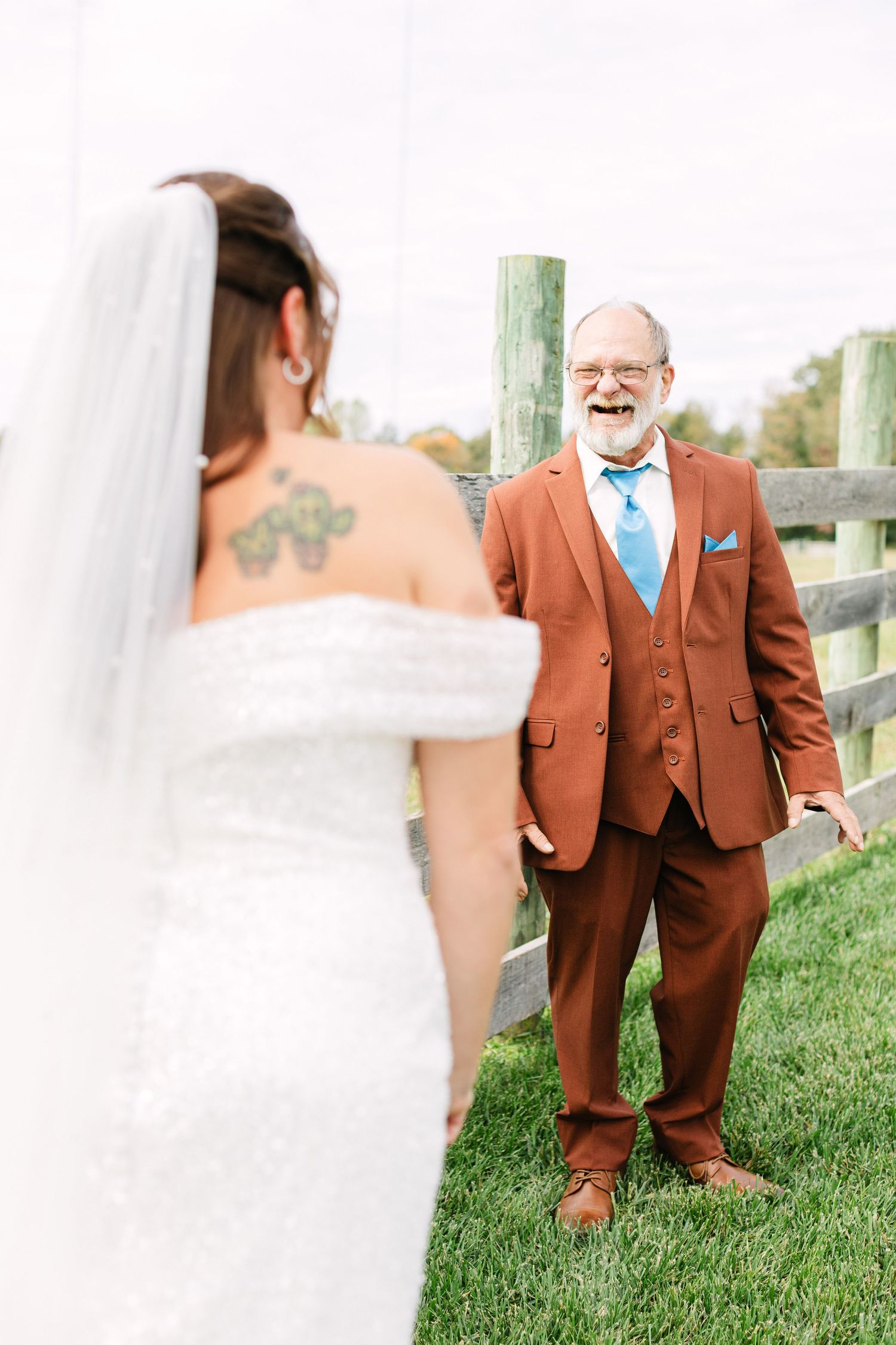 Bride in white off-shoulder gown with tattoo stands before older man in rust suit near wooden fence at outdoor wedding.