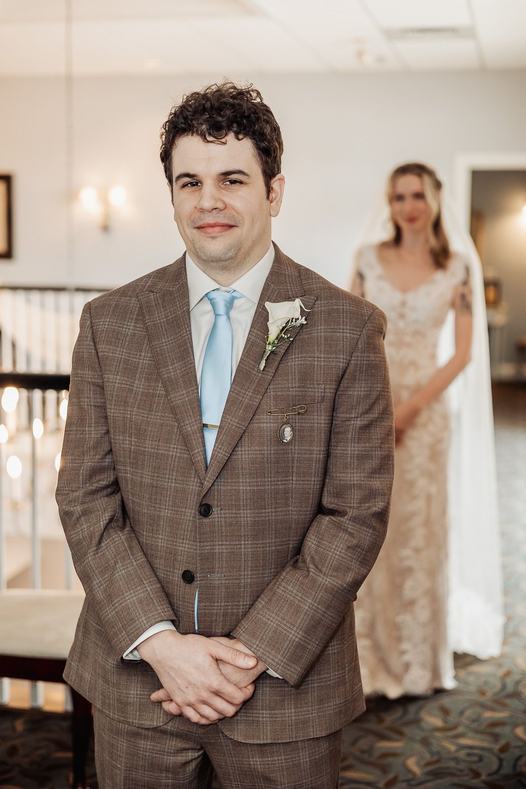 Groom in brown plaid suit smiles while bride in lace gown stands softly blurred in the background.