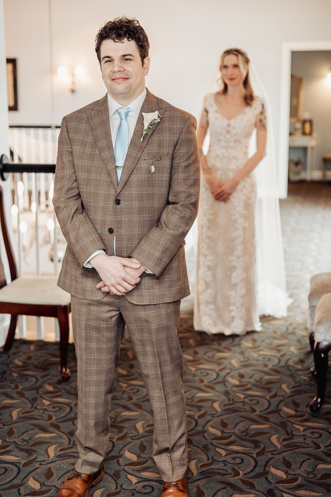 Groom in brown plaid suit with blue tie stands as bride in lace wedding dress waits behind him indoors.