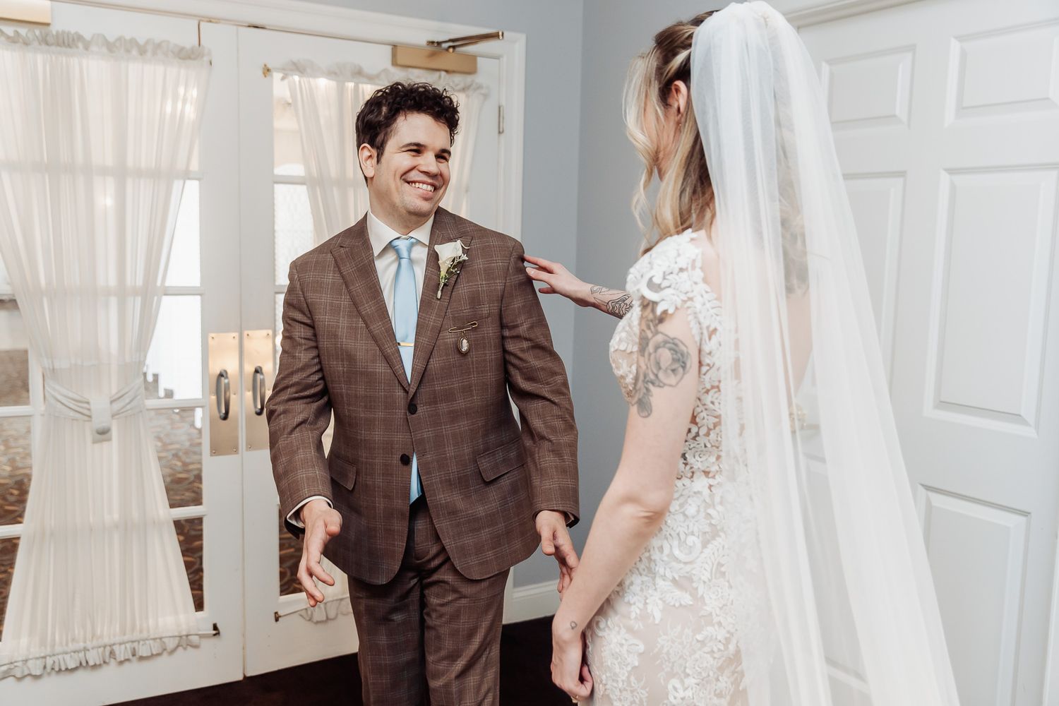 Groom in brown plaid suit smiles at bride in lace wedding dress and veil during first look moment.
