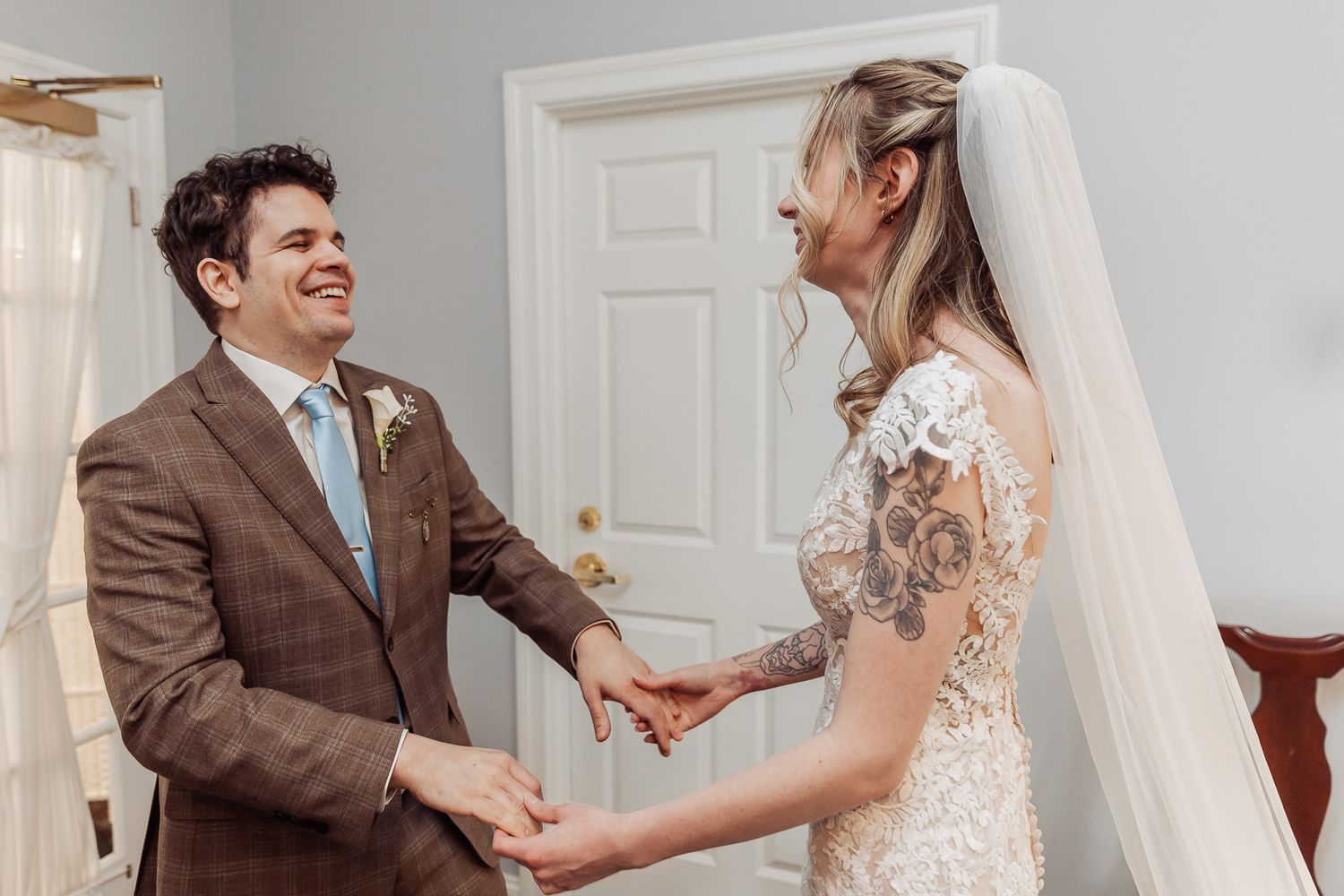 Bride and groom share a joyful first look moment, holding hands and smiling in a sunlit room on their wedding day.