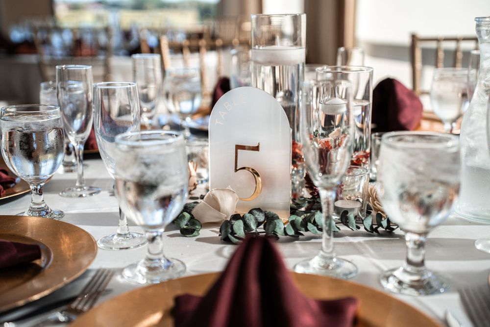 Elegant wedding reception table setting with gold chargers, burgundy napkins, crystal glasses, and a white number 5 centerpiece.