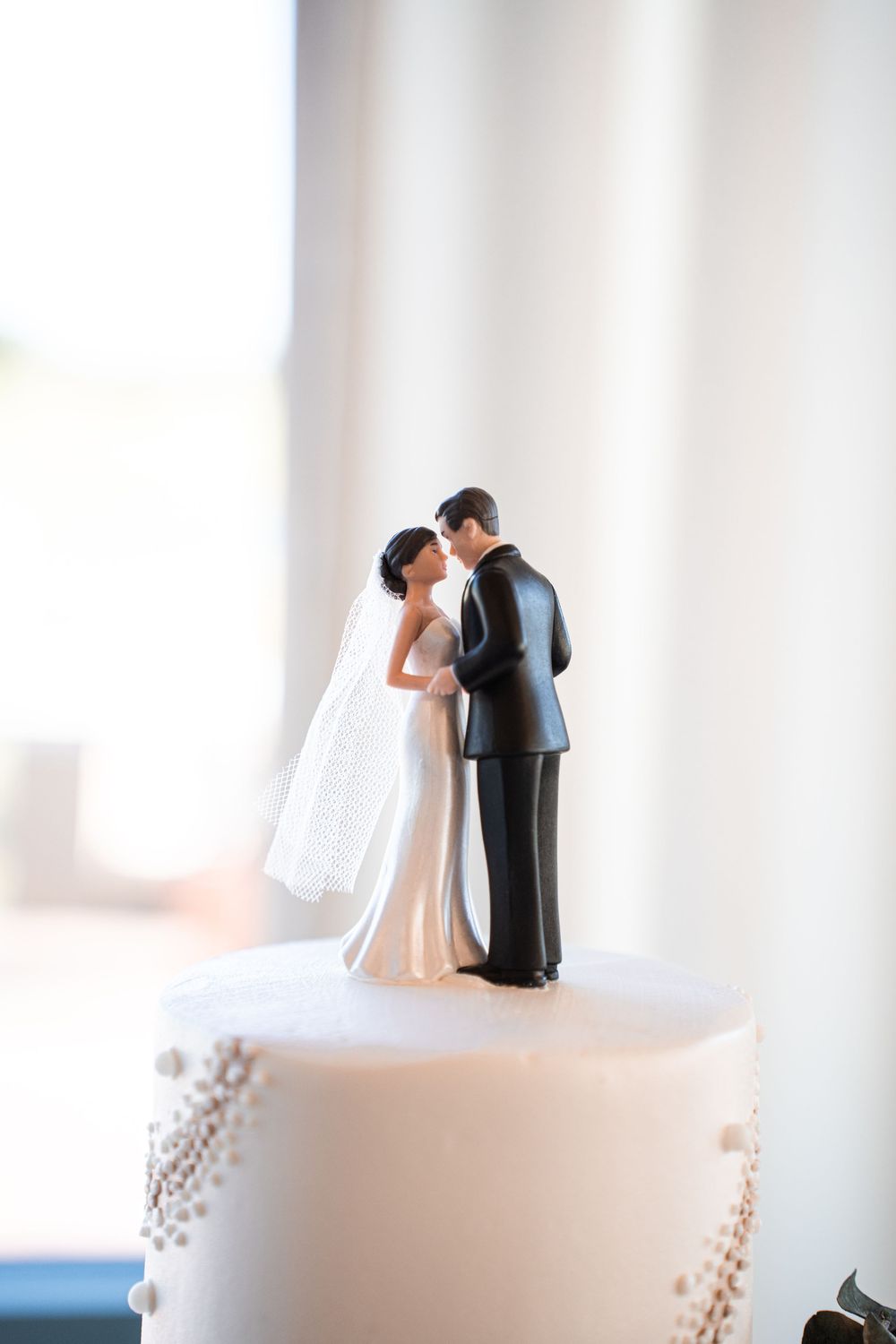Wedding cake topper featuring bride and groom figurines kissing atop a white pearl-decorated cake.