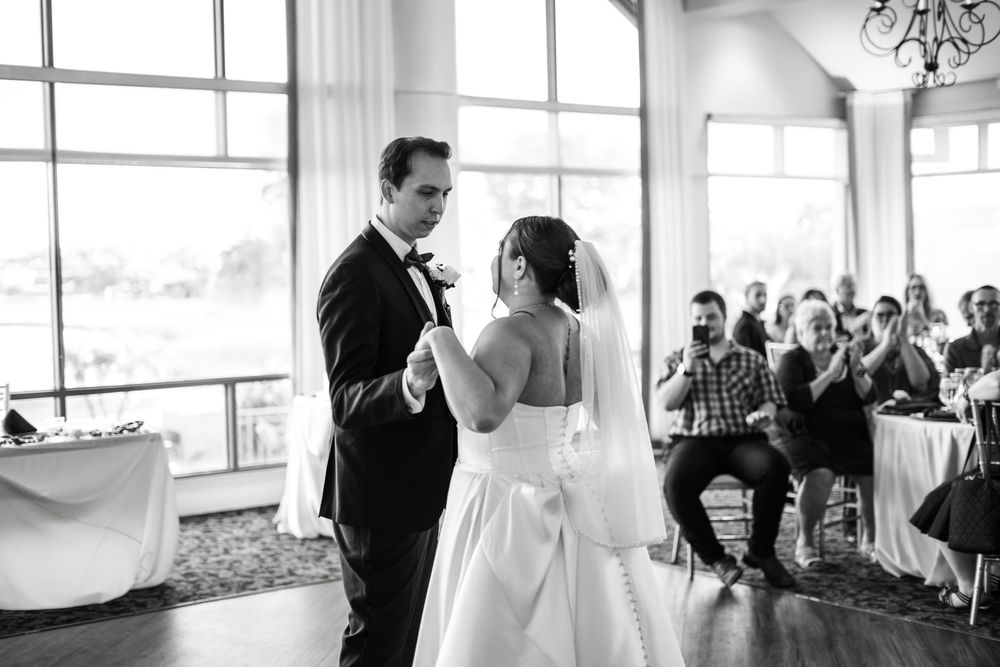Black and white photo of bride and groom sharing first dance at wedding reception while guests applaud.
