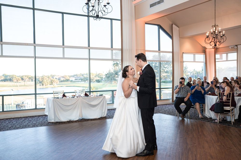 Bride and groom share their first dance in an elegant ballroom with large windows and chandelier.